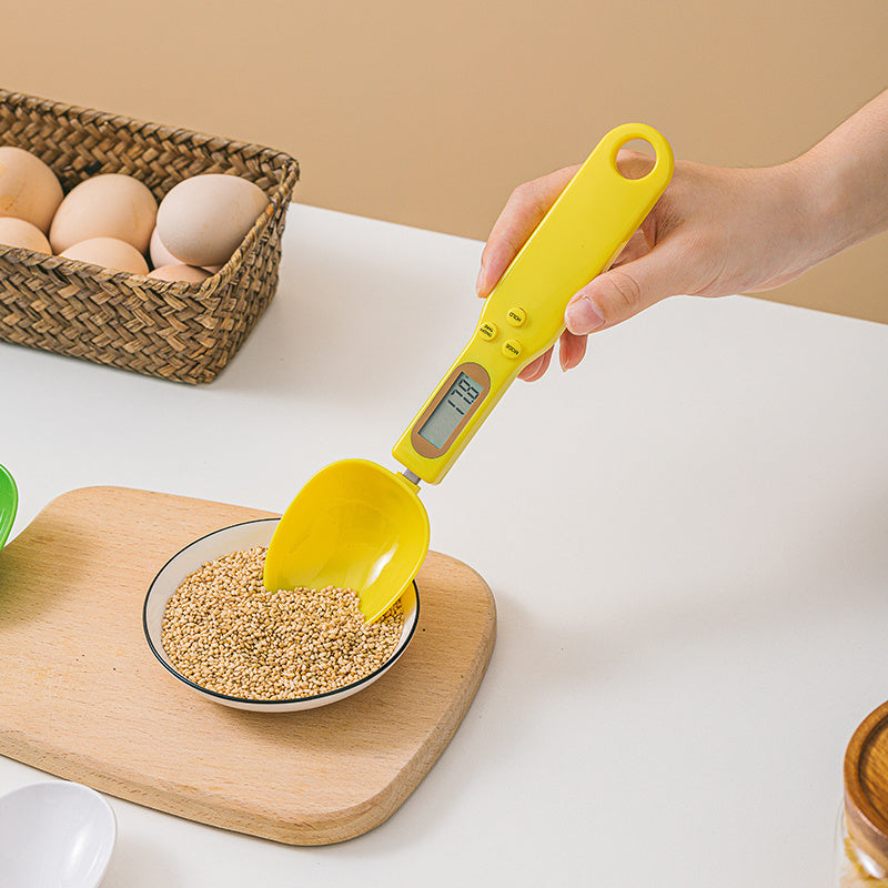 Yellow digital kitchen scoop being used to measure grains on a white surface with a basket of eggs in the background.
